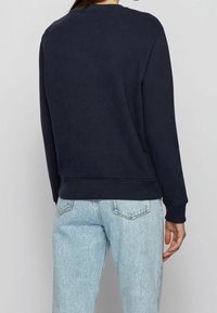 Person som bär en marinblå långärmad sweatshirt och ljusblå denimjeans, sedd bakifrån mot en enfärgad bakgrund.
