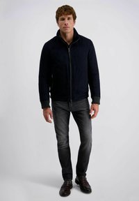 Marineblauwe suède jas met hoge kraag, voorrits en geribbeld detail bij de mouwen, gecombineerd met grijze jeans en donkere schoenen.