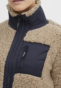 Veste doublée en fleece de couleur beige avec une fermeture éclair navy à l'avant, un tissu texturé et un design à col haut.