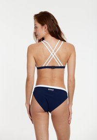 Mornarska bikini postava z belimi poudarki. Zgornji del ima čezkrižne naramnice in zaponko na hrbtu. Spodnji del ima bel pas.