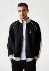 Veste zippée noire avec des accents crème et des manches côtelées, arborant un petit logo sur la poitrine et un col haut.