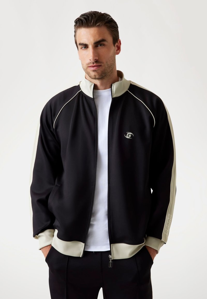 Veste zippée noire avec des accents crème et des manches côtelées, arborant un petit logo sur la poitrine et un col haut.