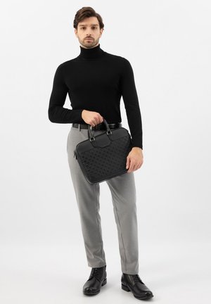 Man in zwarte coltrui, grijze broek en zwarte laarzen die een zwarte textuurkoffer vasthoudt tegen een effen witte achtergrond.