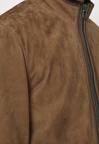 Brun mockaskinnjacka med dragkedja, ribbad krage och strukturerad yta. Har subtila tonala variationer och kontrasterande sömmar.