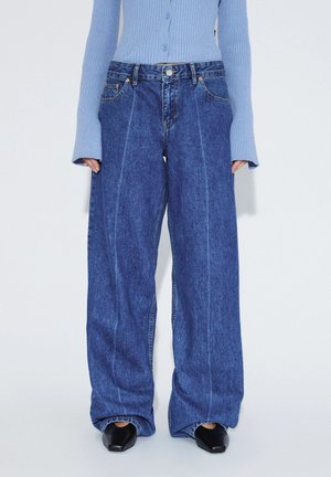 Kvinde iført vidde blå jeans med markante forreste sømme, kombineret med en ribbet lyseblå knaptrøje og sorte spidse sko.