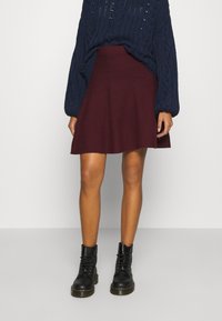 Jupe évasée bordeaux en tissu doux, associée à des bottines noires à lacets et à un pull en tricot torsadé bleu marine.