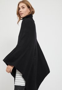 Poncho preto de gola alta com um design solto e fluido. Tecido suave e texturizado que cai até uma bainha pontiaguda. Camisa às riscas visível por baixo.