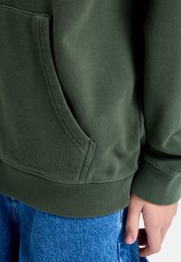 Grön sweatshirttyg med ribbad fåll och manschett, med en sidoficka och en slät yta. Kombinerad med blå denim.