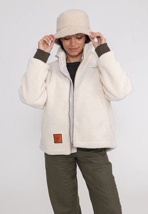 Femme ajustant un chapeau bob crème, portant une veste polaire crème et un pantalon vert olive sur fond uni.