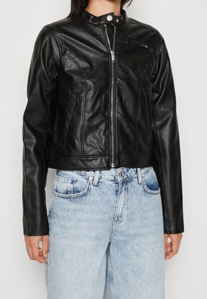 Kobieta ubrana w czarną kurtkę ze sztucznej skóry na suwak z kołnierzem na zatrzaski i jasnoniebieskie dżinsy z wysokim stanem, stojąca na białym tle.