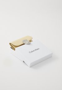Calvin Klein odinė piniginė šviesiai geltonos spalvos, su tekstūruotu paviršiumi ir sidabro spalvos užsegimu, pateikta baltame prekės ženklo dėžutėje.