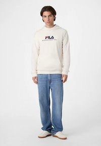 Krämfärgad hoodie med "Fila"-logotyp i marinblått och rött, kombinerad med ljusblå jeans och vita sneakers. Enkel design, avslappnad passform.