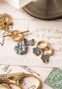 Auksinio papuošalų komplektas su gėlių ir drugelio dizainu. Mėlyno emalio akcentai su subtilia tekstūra. Apima kaklo papuo�šalą ir auskarus.