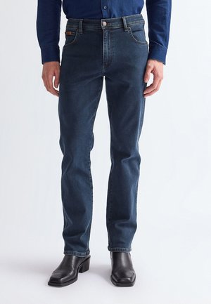 Mand iført mørkeblå slim-fit jeans, navy skjorte og sorte læderstøvler, stående med arme afslappet ned langs siden mod hvid baggrund.