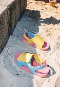 Bunte pinke, gelbe und blaue Kindersandalen auf sandigem Boden nahe einem Steinabsatz, mit einem Strand-Eimer im Hintergrund.