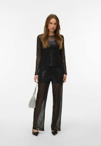 Mustast läbipaistvast riidest komplekt tekstuurse teemantmustriga; pikkade varrukatega top, mida kannavad laia säärega püksid; metallik käekott; elegantsed teravnina kingad.