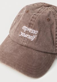 Καφέ βαμβακερό καπέλο μπέιζμπολ με λευκό κεντητό κείμενο "espresso yourself" στο εμπρόσθιο πάνελ, τοποθετημένο σε λευκή επιφάνεια.