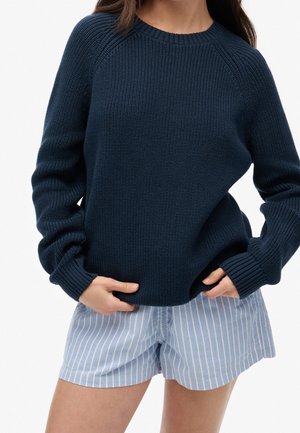 Kobieta ubrana w granatowy sweter z dzianiny z prążkowanymi mankietami oraz jasnoniebieskie i białe pionowo paskowane krótkie spodenki, dłonie wsunięte pod brzeg swetra.