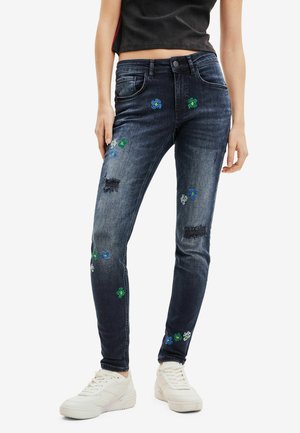 Jeans in denim blu scuro skinny-fit con ricami floreali blu e verdi, zone sbiadite e stile a cinque tasche. Indossati con sneakers bianche.