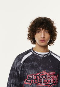 Črna in siva grafična majica s kratkimi rokavi, ki ima bele dodatke in rdeče besedilo. Tkanina ima teksturiran vzorec, z okroglo ovratnico in raglan rokavi.