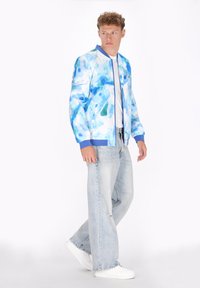 Blauw-witte bomberjack met abstract aquarelpatroon, contrasterende blauwe kraag, gecombineerd met lichte denim jeans en witte sneakers.
