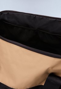 Intérieur d'un sac en tissu beige avec une doublure noire. Dispose d'une fermeture éclair en haut et de poches en maille à l'intérieur pour une meilleure organisation. Texture lisse, design simple.