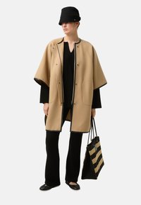 Beige kappefrakke med sort kant, trykknaplukninger, to sidelommer; kombineret med sort top, vidde bukser og en stribet taske. Sort bøllehat.