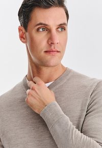 Beżowy sweter z dekoltem w serek, dzianinową fakturą i ściągaczami na mankietach; osoba delikatnie dotyka dekoltu, na jasnym tle.