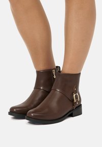 Botas de tobillo de cuero marrón con un dise�ño elegante, que presentan una cremallera lateral y un acento de hebilla en tono dorado en el tobillo. Suela plana negra.