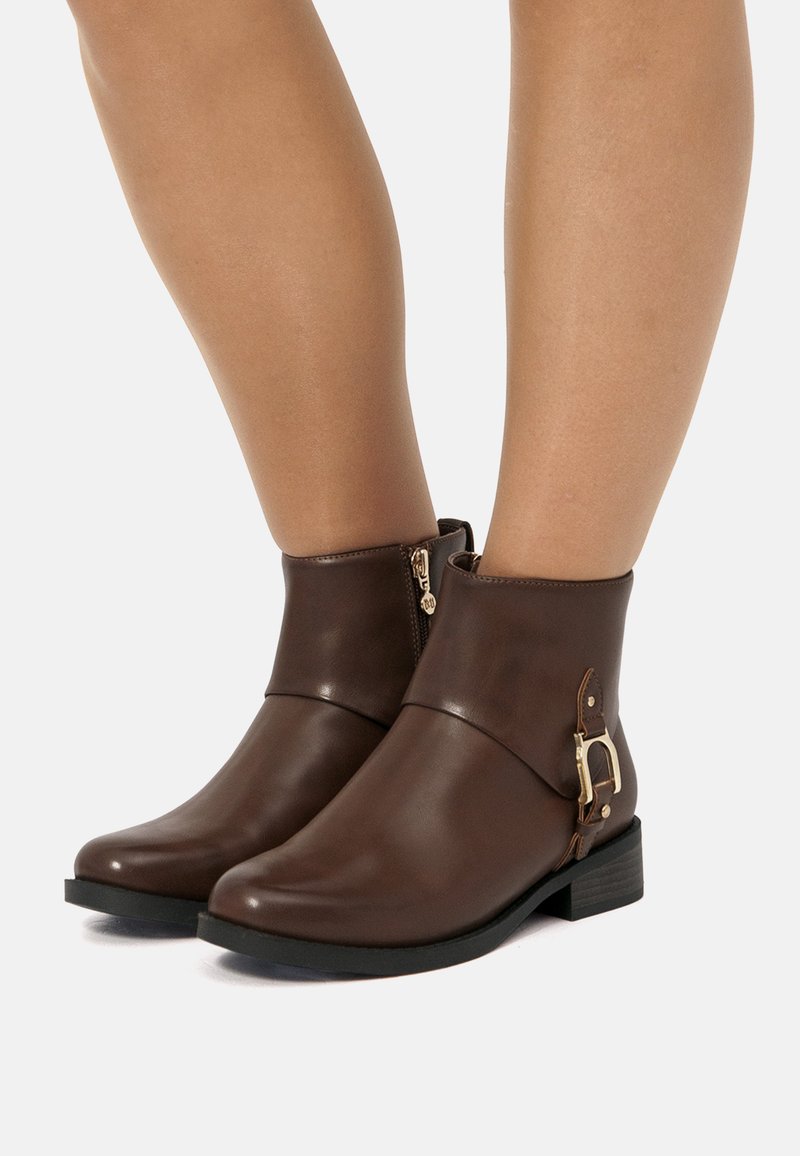 Botas de tobillo de cuero marrón con un diseño elegante, que presentan una cremallera lateral y un acento de hebilla en tono dorado en el tobillo. Suela plana negra.