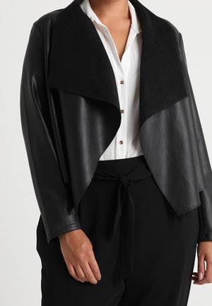Femme portant une veste en cuir noire à larges revers sur une chemise blanche boutonnée et un pantalon noir taille haute avec une ceinture nouée.