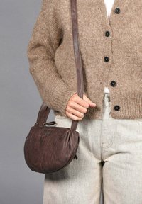 Bolso de cuero marrón tipo bandolera con forma redondeada, superficie texturizada, correa corta y un pequeño detalle de logo. Combinado con un cárdigan beige.