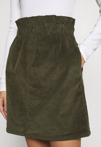 Vrouw in een olijfgroene corduroy rok met een paperbag taille en zijzakken, gecombineerd met een witte blouse met lange mouwen, hand in de zak.