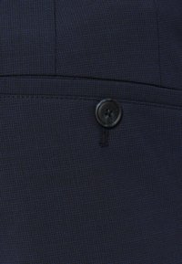 Ναυτικό μπλε υφαντό ύφασμα με διακριτό σχέδιο, διαθέτοντας ένα μόνο μαύρο κουμπί και μια ραμμένη γραμμή πάνω από τη λεπτομέρεια του κουμπιού.