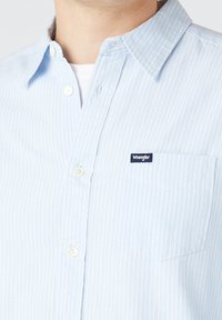 Camicia azzurro chiaro a righe con bottoni, realizzata in tessuto morbido, con taschino sul petto e piccolo logo del marchio sul taschino.