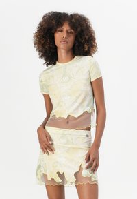 Kortermet tettsittende topp og miniskjørt i gjennomsiktig gul stoff med blomsterdesign, bølgede kanter og kontrasterende skjellkant, med et logo aksent.