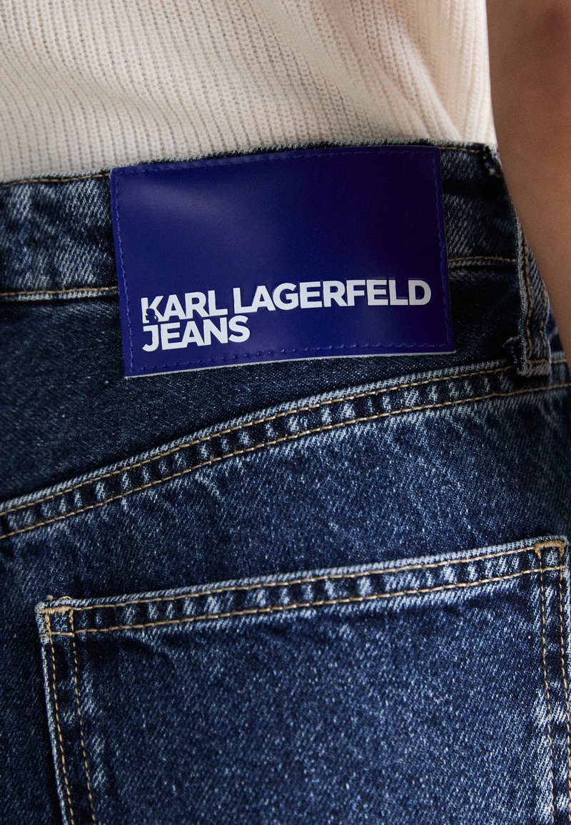Ausgebleichte blaue Denim-Jeans mit einem sichtbaren Patch, der "KARL LAGERFELD JEANS" in fettweißer Schrift auf einem rechteckigen lila Hintergrund zeigt.