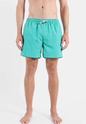 Homme debout pieds nus portant un short de bain turquoise avec un cordon blanc sur un fond clair uni, vue de face de la taille vers le bas.