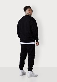 Schwarzer Pullover und Jogginghose mit gerippten Bündchen, kombiniert mit weißen Sneakers. Merkmale umfassen einen Lagenlook mit einem weißen Hemd darunter.