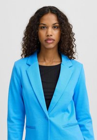 Blauwe blazer met een gladde textuur, met notch-revers, knoopsluiting en twee voorkanten zakken, gedragen over een zwart geribbeld top.