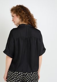 Chemise noire à manches courtes avec col, coupe décontractée et détail plissé au dos, associée à un pantalon à motif à pois. Texture de tissu douce.