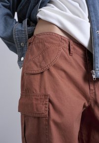 Κοντινό πλάνο ενός ατόμου που φοράει πα ντελόνι cargo σε χρώμα σκουριάς, λευκό πουκάμισο μέσα από το παντελόνι και ένα μερικώς ανοιχτό μπλε τζιν μπουφάν.