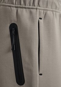 Beige Hose aus strukturiertem Stoff, mit einer schwarzen Reißverschlusstasche an der Seite und einem elastischen Bund. Dezente Nahtdetails sichtbar.