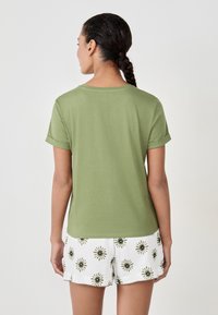 Frau mit geflochtenem Haar, die steht und ein olivgrünes Kurzarmshirt sowie weiße Shorts mit grünen Blumenmustern trägt.