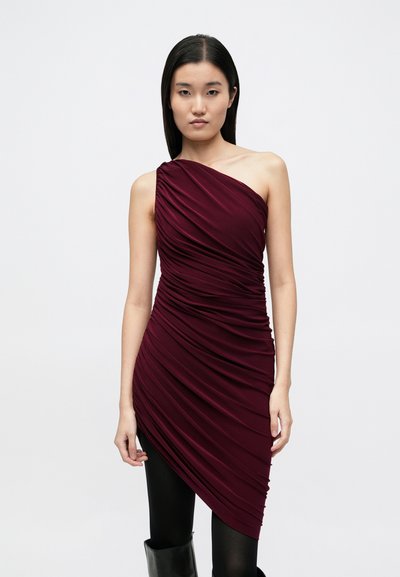Ein schulterfreies burgunderrotes Kleid mit geraffter Textur, asymmetrischem Saum und schlanker Passform. Das Modell trägt schwarze Leggings und kniehohe Stiefel.