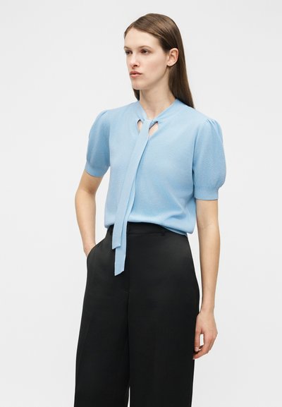 Femme portant un haut à manches courtes bleu clair avec un col noué et un pantalon large noir taille haute, debout avec une main dans la poche.