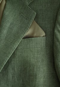 Grüne Leinenanzugjacke mit einem glatten Innenfutter, ausgestattet mit einer Pattentasche und dezenten Nähdetails am Revers.