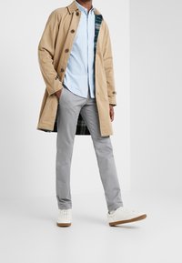 Beige trenchcoat med knappar, blå skjorta, grå byxor och vita sneakers med bruna sulor; rutigt foder synligt i kappan.