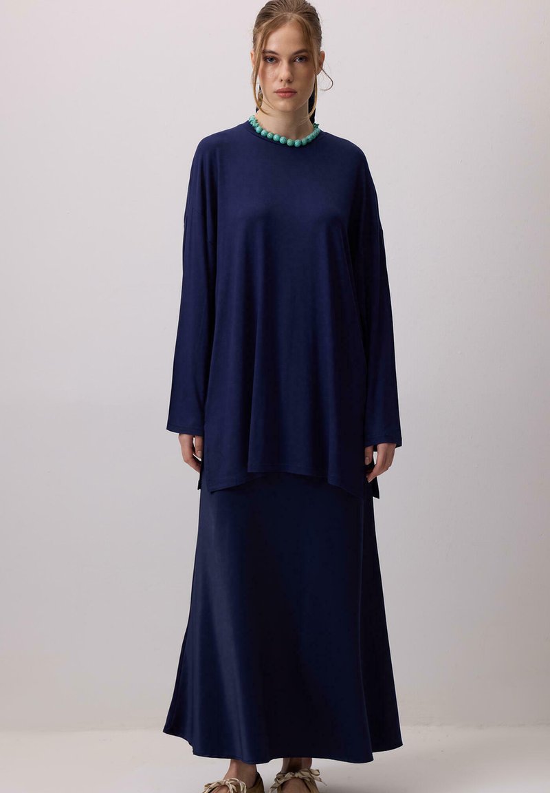 Γυναίκα που φοράει χαλαρή μπλε σκούρα μπλούζα με μακριά μανίκια και μακριά φούστα στο ίδιο χρώμα, με τυρκουάζ κολιέ από χάντρες, στέκεται μπροστά σε απλό ανοιχτό φόντο.