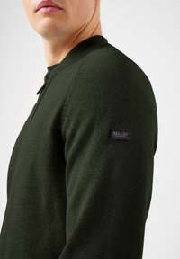 Mężczyzna w ciemnozielonym dzianinowym swetrze zapinanym na zamek, z logo Maerz München na górnym rękawie, na białym tle.